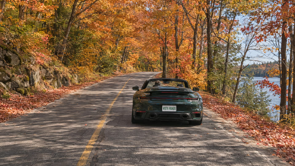 A green convertible on a two-lane road under red and gold maples in fall