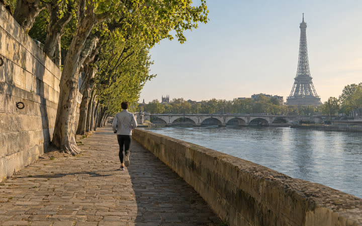 A stone riverside path under plane trees, with a river and a tall iron tower ahead