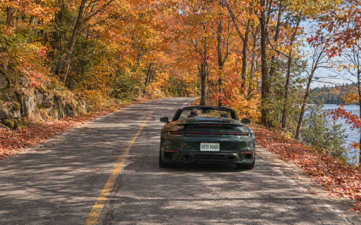 A two-lane road under red and gold maples, with a lake through the trees